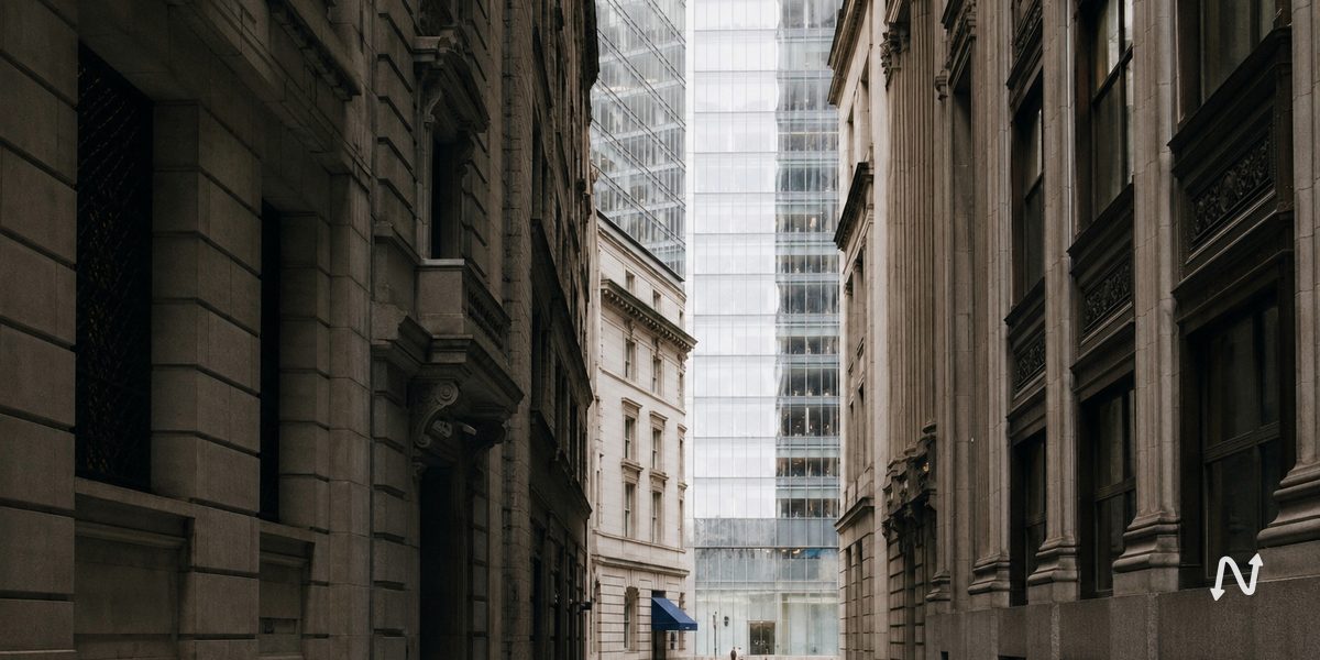 A narrow street between older stone commercial buildings opening onto a modern glass tower, under flat overcast light.